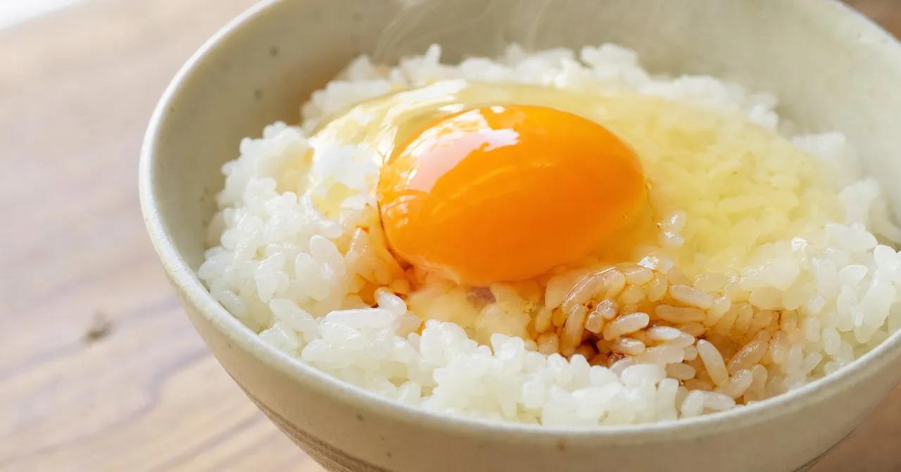 A bowl of hot white rice topped with a raw egg yolk and a little soy sauce, steam rising, on a plain wooden table.