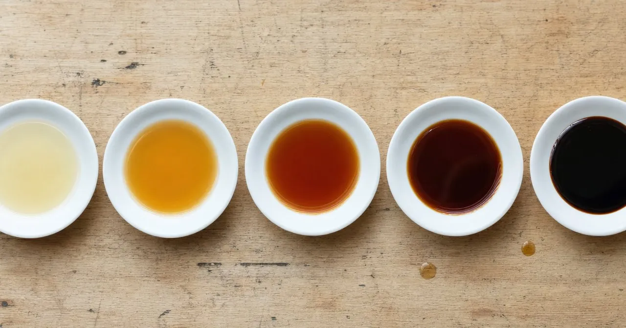 Five small saucers of Japanese soy sauce in a row, from pale gold shiro and amber usukuchi to red-brown koikuchi, dark tamari and nearly black saishikomi.
