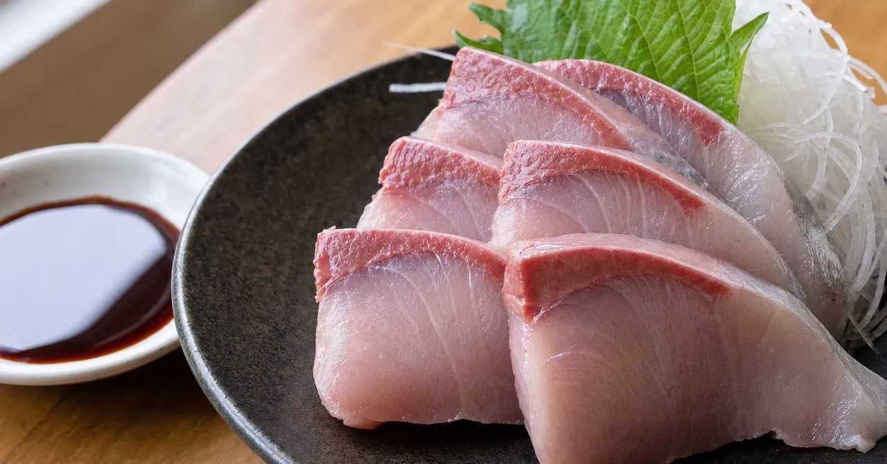 A plate of glossy yellowtail sashimi with shredded daikon and a shiso leaf, next to a small dish of soy sauce.