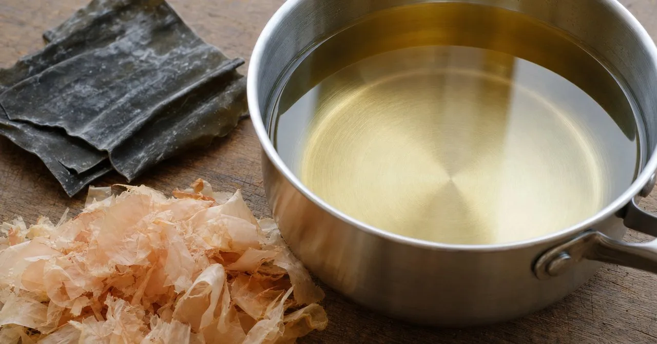 A small saucepan of clear, pale golden dashi with a piece of kombu and a soft heap of katsuobushi flakes beside it on a wooden counter.